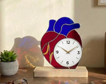 May include: A heart-shaped clock made of stained glass, with red and blue segments and a white clock face with gold hands. It sits on a wooden base. The brand name "Lightinggo" is visible in the top left corner.