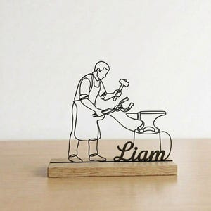 May include: Black metal blacksmith sculpture on a wooden base. The sculpture depicts a blacksmith working with a hammer and tongs near an anvil. The name "Liam" is written in cursive script below the blacksmith.