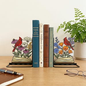 May include: A pair of black metal bookends with a floral and cardinal design. The bookends hold several books, including "Wildlife of North America" and "Nature's Designs." A pen, notebook, and glasses are on the wooden surface.