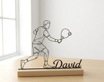 May include: A black metal tennis player figurine with the name "David" on a wooden base. The figurine shows a tennis player in action, holding a racket and ball. The design is a minimalist line drawing.