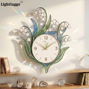 May include: A wall clock featuring a white circular face with gold hands and a lily of the valley illustration. The clock is surrounded by a decorative stained glass frame in shades of green and white, shaped like lily of the valley flowers.
