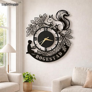 May include: Black metal wall clock featuring a squirrel perched on a branch with acorns and leaves. The clock face is black with gold hands. A silhouette of a child is on the left, and the name "BOGESTAD" is at the bottom.