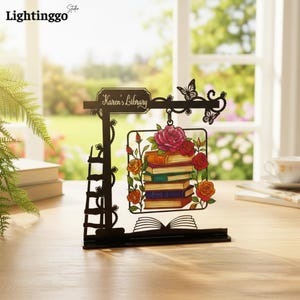 May include: A black decorative sign featuring the text Karen's Library, a ladder, and butterflies. A central square frame displays a stack of colourful books surrounded by red, pink, and orange roses, all resting on a base shaped like an open book.