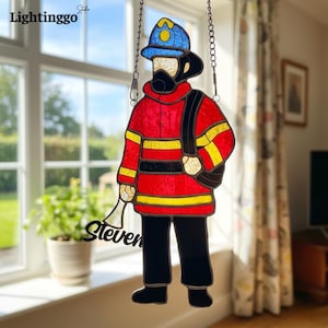 Puede incluir: Figura de bombero de vidriera con chaqueta roja, pantalones negros y casco azul. La figura tiene rayas amarillas y una bolsa negra. El nombre "Steven" está escrito en cursiva negra. La imagen cuelga de una cadena.