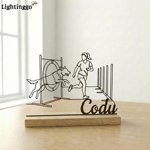 Könnte beinhalten: Eine Hundesport-Szene aus schwarzem Metall mit einem Hund, der durch einen Reifen springt, einer Person, die rennt, und dem Namen "Cody" auf einem Holzsockel. Das Design ist im Strichstil gehalten.