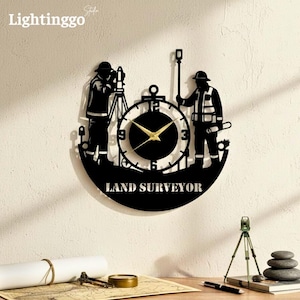 Op de afbeelding: Zwarte metalen wandklok met een landmeterontwerp. De klok heeft een ronde wijzerplaat met gouden wijzers en de tekst "LAND SURVEYOR" onderaan. Aan weerszijden van de klok staan twee landmeterfiguren.
