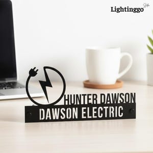 May include: Black metal desk sign with the text "HUNTER DAWSON" and "DAWSON ELECTRIC" in white. The sign features a lightning bolt and plug symbol. A white mug and laptop are in the background.