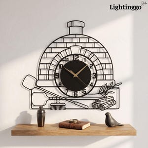 Puede incluir: Un reloj de pared de metal negro con forma de horno de pizza de ladrillo con chimenea. La esfera es circular con manecillas doradas y números negros. Debajo del diseño del horno hay siluetas de una pala, un cepillo y leña.