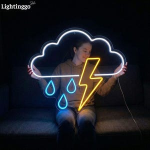 May include: A neon sign in the shape of a cloud with a lightning bolt and raindrops. The cloud outline and raindrops are blue, and the lightning bolt is yellow. The sign is held by a person.