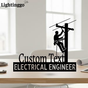 Puede incluir: Letrero de metal negro con la silueta de un ingeniero eléctrico trabajando en un poste de energía. El letrero incluye el texto "Custom Text" encima de "ELECTRICAL ENGINEER". El logotipo de "Lightinggo" está en la esquina superior izquierda.
