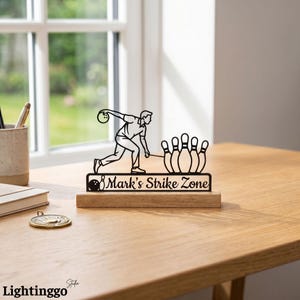 May include: A black metal bowling-themed decorative piece featuring a bowler, pins, and the text "Mark's Strike Zone" on a wooden base. The design is set against a window background.