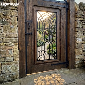 May include: A dark brown wooden gate featuring a central black metal insert with an arched, repeating geometric pattern. The gate is mounted with black iron hinges and a handle, set within a rustic stone wall with a stone path visible through it.