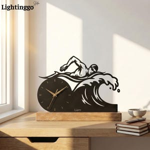 Könnte beinhalten: Eine Tischuhr aus schwarzem Metall mit der Silhouette eines Schwimmers in einer Welle. Das Zifferblatt ist kreisförmig mit goldenen Zeigern und der Name Liam ist an der Basis aufgedruckt. Die Uhr steht auf einem rechteckigen hellen Holzsockel.