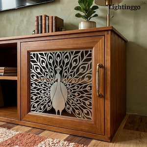 May include: A wooden cabinet featuring a single door with a silver-toned metal panel displaying a laser-cut peacock design. The cabinet has a vertical brass-colored handle, an open side shelf with stacked books, and a smooth, polished wood finish.