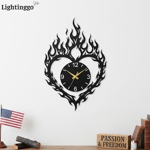 May include: Black wall clock with a flame-shaped frame and a round clock face. The clock has gold hands and a black background. The clock is surrounded by a flame design. The clock is mounted on a white wall.