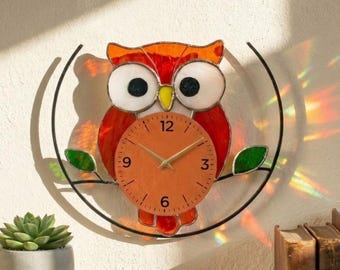 May include: A stained glass owl wall clock with an orange clock face. The owl features red and orange feathers, large white eyes, and a yellow beak. The clock is mounted on a black metal frame with a branch and green leaves. The text "Lightinggo" is in the top left corner.