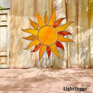 May include: A sun-shaped stained glass suncatcher featuring a central circular yellow piece surrounded by alternating pointed rays in shades of orange and deep red, all held together by thin dark metal solder lines.
