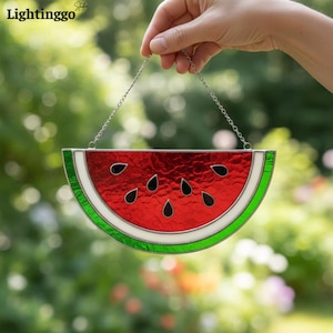 May include: A stained glass watermelon slice suncatcher with a silver chain for hanging. The watermelon features red, green, white, and black details. The suncatcher is held up by a hand against a blurred green background.