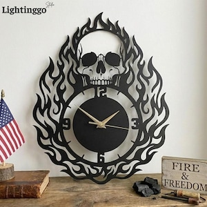 Könnte beinhalten: Schwarze Metall-Wanduhr mit einem Totenkopf-Design, umgeben von Flammen. Das Zifferblatt hat goldfarbene Zeiger und ein minimalistisches Design. Die Uhr ist an einer weißen Wand montiert, mit einer kleinen amerikanischen Flagge und einem Holzschild mit "FIRE & FREEDOM".