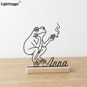 May include: A black metal frog sculpture, holding a cigarette and exhaling smoke, is mounted on a wooden base. The name "Anna" is written in cursive below the frog. The frog is in a relaxed pose, with its chin resting on its hand.