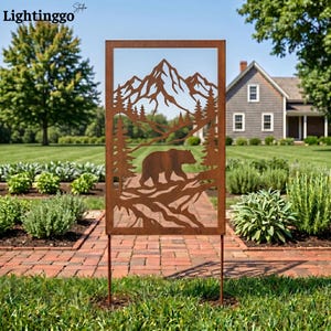 May include: A rectangular, rusty-brown metal garden art piece featuring a bear, mountains, and trees silhouette. The artwork is mounted on two stakes and is displayed outdoors in a garden setting.