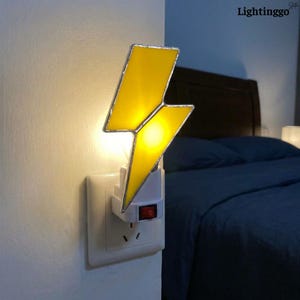 Può includere: Una luce notturna a forma di fulmine giallo collegata a una presa a muro bianca. La luce notturna ha un bordo argentato e un interruttore on/off rosso. Lo sfondo include un letto blu scuro e una testiera marrone.