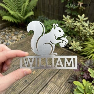 Puede incluir: Silueta metálica plateada de una ardilla sosteniendo una bellota, con el nombre "WILLIAM" recortado debajo. La ardilla tiene un acabado texturizado. El fondo presenta vegetación y una superficie de madera.