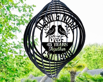 May include: Black metal wind spinner with the text "ALAN & NOAH" and "45 YEARS Together EST. 1980". The design includes two birds perched on a branch. The wind spinner is hanging outdoors.