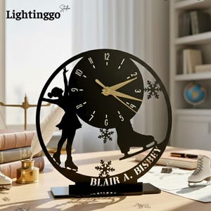 May include: Black decorative clock featuring a figure skater silhouette and a clock face. The clock has gold-colored hands and the name "BLAIR A. BISBEY" is displayed at the bottom. The clock is on a wooden surface.
