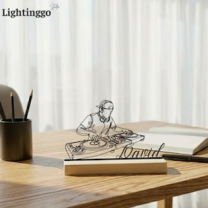 Puede incluir: Una representación en línea negra de un DJ con auriculares y tocadiscos, montada sobre una base de madera. El nombre "David" está escrito en cursiva. El logotipo "Lightinggo" está en la esquina superior izquierda.