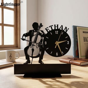May include: A black desk clock featuring a silhouette of a musician playing a cello. The circular clock face displays the name ETHAN at the top, with white numbers 3, 6, 9, and 12, and gold-colored hour, minute, and second hands on a stand.