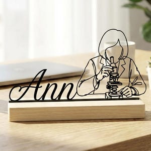 Peut inclure: Un porte-nom de bureau noir découpé au laser avec le nom "Ann" en écriture cursive. Le porte-nom présente un dessin au trait d'une personne utilisant un microscope. La base est en bois de couleur claire.