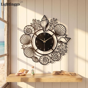 May include: A circular wall clock featuring a black face with gold hands and numerals at 3, 6, 9, and 12. The frame is a black metal cutout design depicting various seashells and coral branches arranged in a decorative wreath pattern around the clock.
