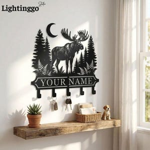 May include: Black metal wall-mounted key holder featuring a moose silhouette, trees, and a crescent moon. The key holder has hooks below a banner that reads "YOUR NAME". A small potted plant, keys, and a basket sit on a wooden shelf below.