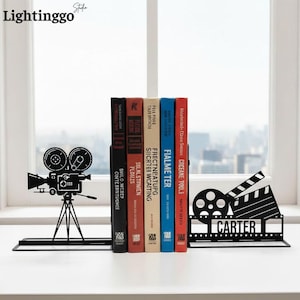 May include: Black metal bookends in a movie theme. One bookend features a film camera silhouette, the other a film reel and clapperboard with the name "CARTER". Books are held between the bookends on a white surface.