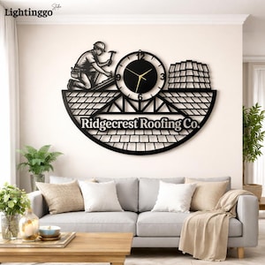 May include: Black metal wall clock depicting a roofer, a clock face, and the text "Ridgecrest Roofing Co." The clock has a roof design. The clock is mounted on a white wall above a grey sofa. The clock hands show the time.