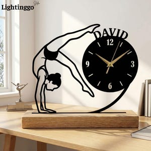 May include: A black metal silhouette of a gymnast performing a backbend, attached to a circular clock face with gold hands and the name DAVID printed in white. The clock is mounted on a rectangular light-colored wooden base.