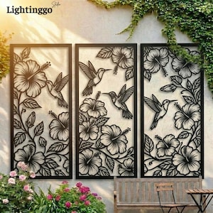 May include: Three-panel black metal wall art featuring hummingbird and hibiscus flower designs. The laser-cut panels are framed and create a decorative outdoor or indoor accent. The panels are mounted on a light-coloured wall with greenery.