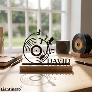 May include: A black metal record player silhouette with musical notes and the name "DAVID" on a wooden base. Two vinyl records and a small speaker are visible in the background. The image is set on a wooden desk.