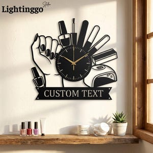 Könnte beinhalten: Eine Wanduhr aus schwarzem Metall mit einer Hand mit manikürten Nägeln, Nagellackflaschen, einer Nagelfeile und einer UV-Lampe. Das Zifferblatt ist rund mit goldenen Zeigern. Ein Banner am unteren Rand zeigt den Text CUSTOM TEXT in Großbuchstaben.