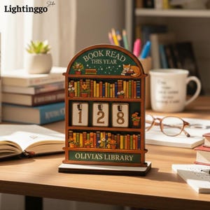 May include: A wooden bookshelf-shaped tracker with the text "BOOK READ THIS YEAR" at the top and "OLIVIA'S LIBRARY" at the bottom. It features three rows of colorful book illustrations, potted plants, a sleeping cat, and three rotating number blocks displaying 128.