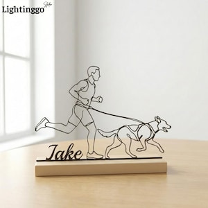 May include: A black metal sculpture depicting a person running with a dog on a leash. The sculpture is mounted on a rectangular wooden base with the name "Jake" in cursive. The Lightinggo logo is in the top left corner.
