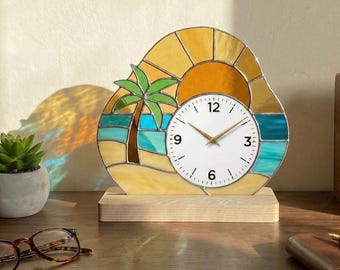 May include: A stained glass clock depicting a beach scene. The clock face is white with black numerals and gold hands. The background features a yellow sun, blue water, a sandy beach, and a green palm tree. The clock sits on a wooden base.