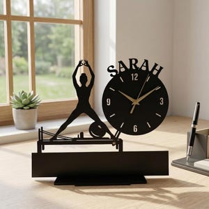 Może przedstawiać: Czarny zegar biurkowy z sylwetką osoby wykonującej ćwiczenie pilates na maszynie typu reformer. Imię SARAH jest wycięte nad okrągłą tarczą zegara, która posiada złote wskazówki oraz białe cyfry 3, 6, 9 i 12 na czarnym tle.