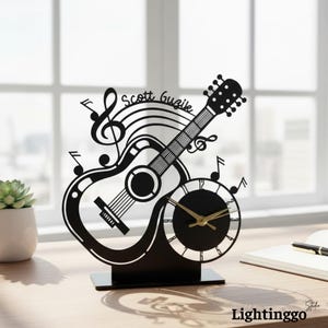 May include: Black guitar-shaped desk clock with musical notes and the name "Scott Guzik" on the fretboard. The clock has gold-colored hands and a round clock face. The clock is on a black base.