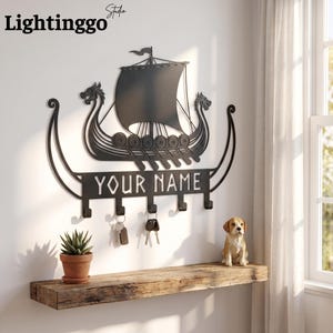 Peut inclure: Un porte-clés mural en métal noir en forme de drakkar viking avec des proues en tête de dragon et une voile. Le design comprend une barre horizontale avec le texte "YOUR NAME" et cinq crochets, avec deux jeux de clés suspendus aux crochets.