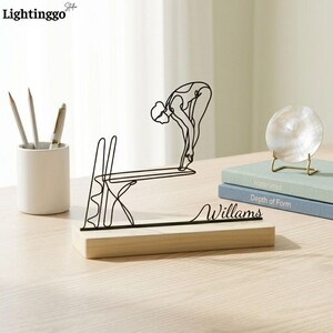 May include: A black metal sculpture of a diver poised to jump, mounted on a light wood base. The sculpture includes the name "Williams" in cursive. The sculpture is a decorative item for a desk or shelf.