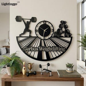 May include: Black metal wall clock with a weightlifting theme. The clock features a silhouette of a weightlifter, a clock face, and a rack of weights. The text "Gerbus Weightlifting" is displayed below the clock. The clock is displayed above a wooden table.