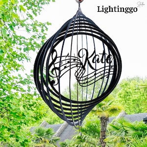 May include: A black metal wind spinner with a spiral design. The spinner features the name "Kate" and musical notes cut out in the center. The background shows green foliage and a house.