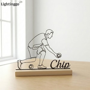 May include: A black line art sculpture of a person playing lawn bowls, with the word "Chip" in cursive. The sculpture is mounted on a rectangular wooden base. The background is a white wall and a wooden table.